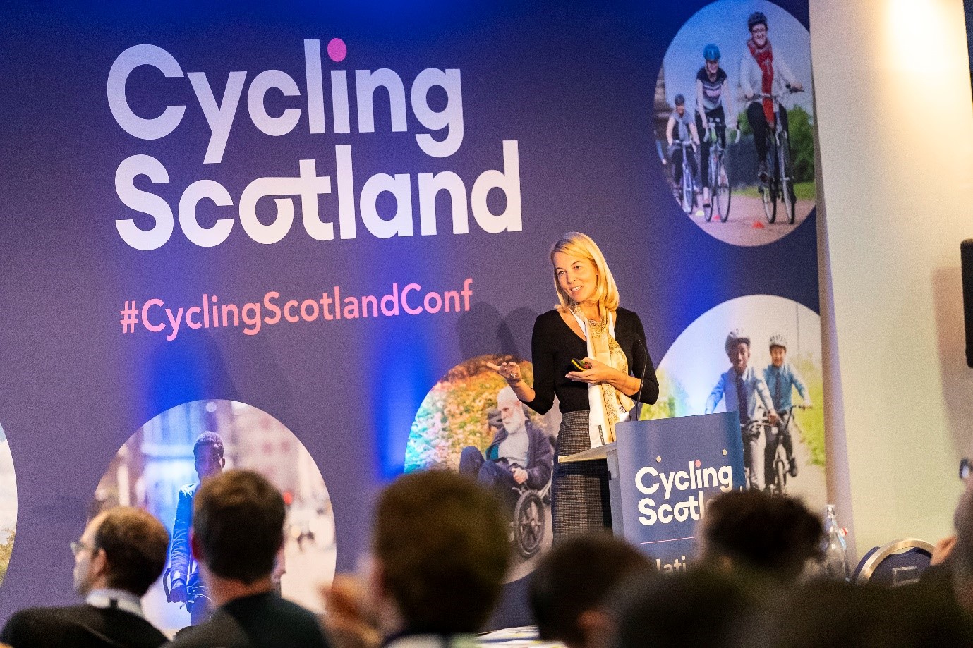 A person at a podium talking to people, with a purple background with white text reading Cycling Scotland