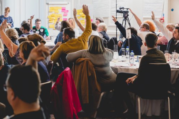 People sit around tables at an event with raised hands to ask questions to a speaker.