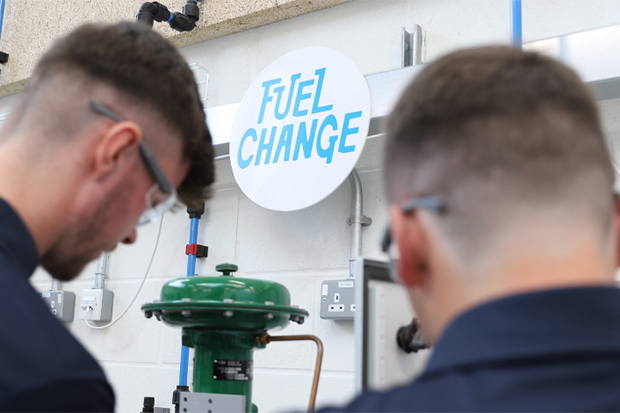 Two engineers working in a workshop with a 'Fuel Change' sign on the wall.