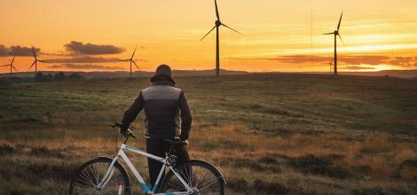 A man leaning against a bicycle looks into the distance at the sunset behind wind turbines