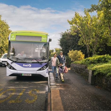 A family walks off a bus onto the pavement of a leafy street.