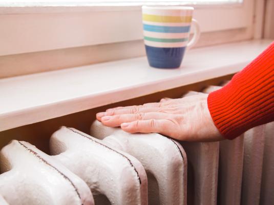 A person in a red jumper placing their hand on a white radiator, with a stripey sitting on the windowsill.