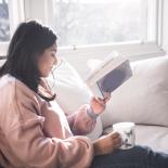 A woman sitting on a couch by a window, reading a book and holding a cup of tea.